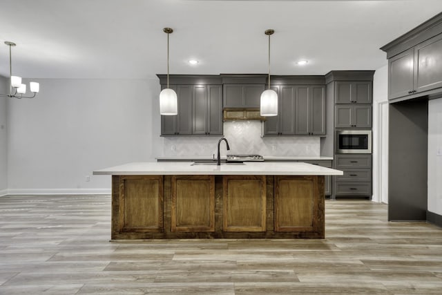 kitchen featuring gray cabinetry, sink, decorative light fixtures, stainless steel microwave, and an island with sink