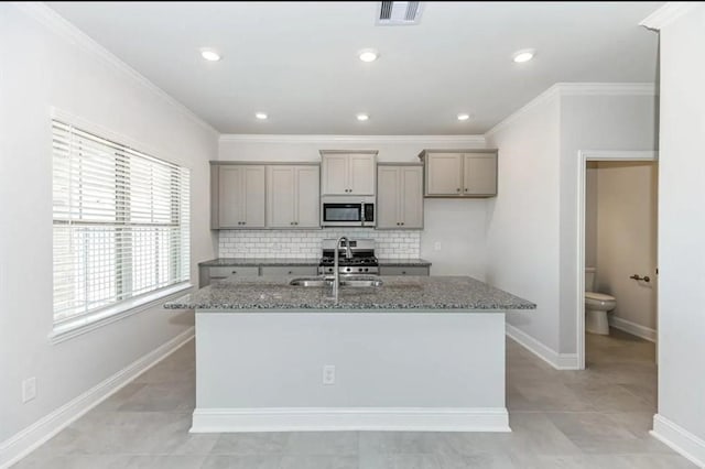 kitchen featuring a kitchen island with sink, crown molding, tasteful backsplash, light stone counters, and appliances with stainless steel finishes