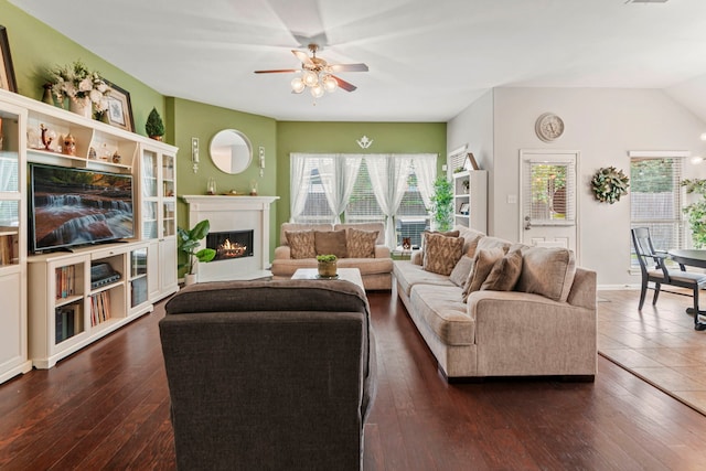 living room featuring baseboards, a warm lit fireplace, dark wood finished floors, and a ceiling fan