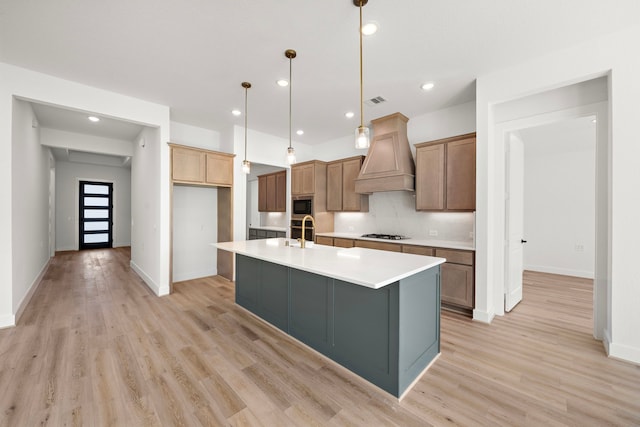 kitchen with gas cooktop, light hardwood / wood-style flooring, premium range hood, decorative light fixtures, and a kitchen island with sink