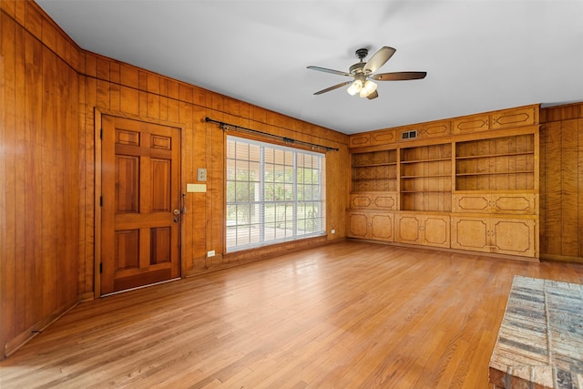 unfurnished living room with built in shelves, ceiling fan, light wood-type flooring, and wooden walls