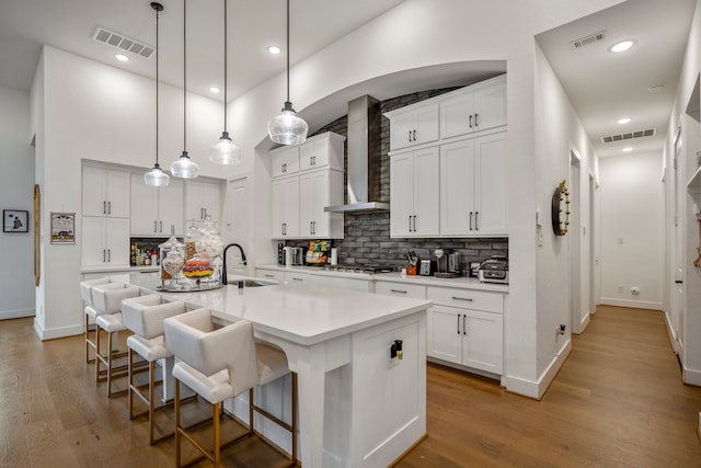 kitchen featuring wall chimney range hood, sink, an island with sink, white cabinetry, and decorative light fixtures