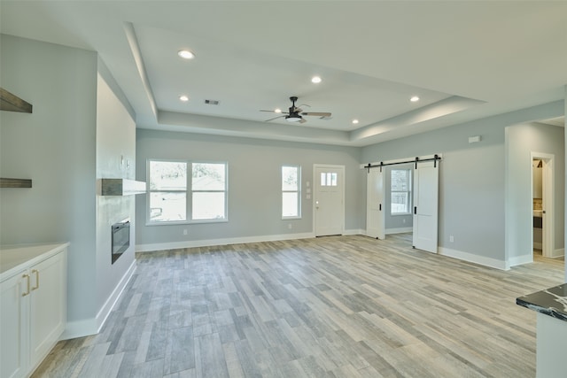 unfurnished living room with a barn door, ceiling fan, a raised ceiling, and light hardwood / wood-style flooring