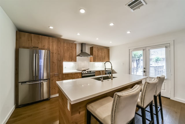 kitchen featuring sink, an island with sink, dark wood-type flooring, appliances with stainless steel finishes, and wall chimney range hood