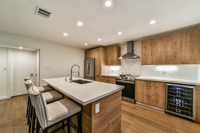 kitchen with stainless steel appliances, sink, wall chimney range hood, an island with sink, and beverage cooler