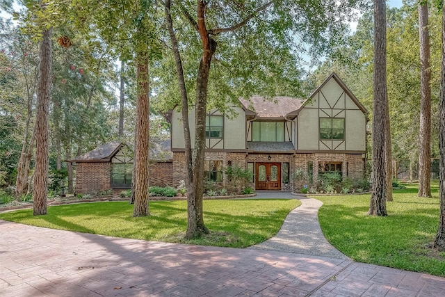 tudor-style house with brick siding, french doors, roof with shingles, stucco siding, and a front yard