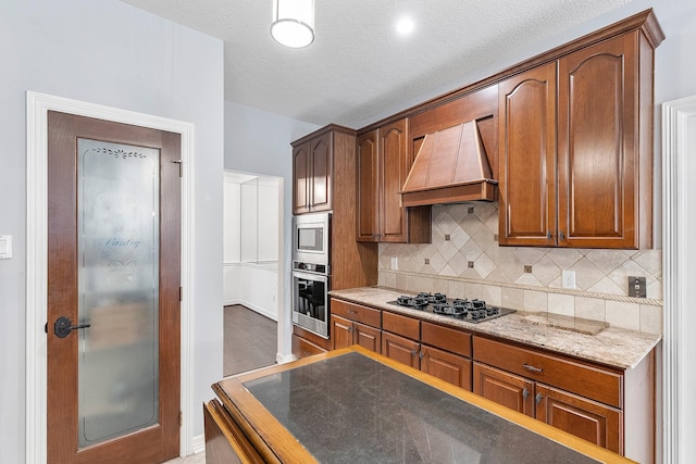 kitchen featuring appliances with stainless steel finishes, premium range hood, tasteful backsplash, light stone countertops, and a textured ceiling