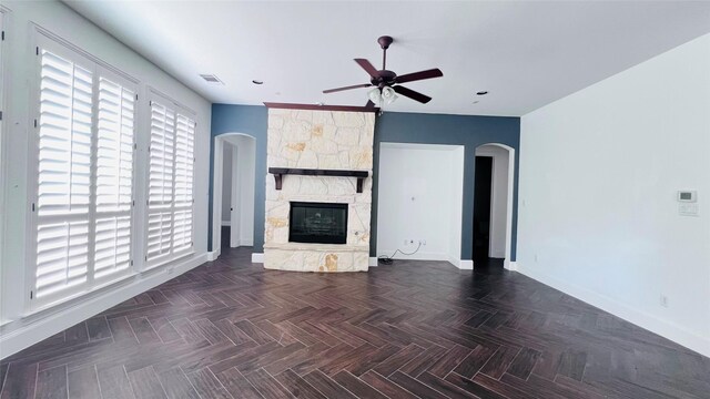 unfurnished living room featuring ceiling fan, a stone fireplace, and dark parquet floors