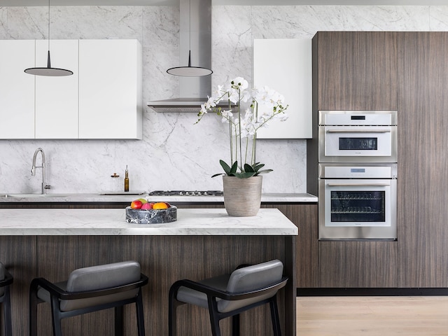 kitchen with multiple ovens, ventilation hood, sink, decorative light fixtures, and a breakfast bar area