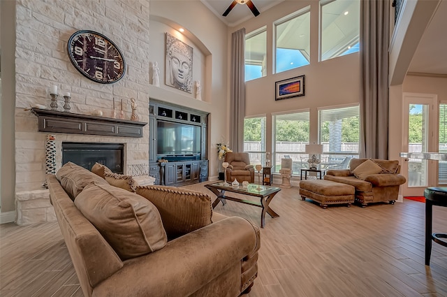 living room featuring light wood-type flooring, ceiling fan, a stone fireplace, a high ceiling, and crown molding