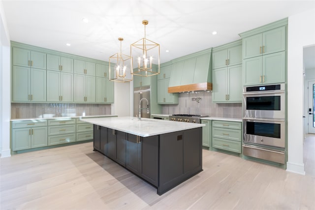 kitchen featuring light stone countertops, appliances with stainless steel finishes, custom range hood, hanging light fixtures, and an island with sink