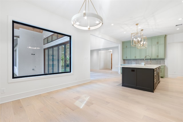 interior space featuring hanging light fixtures, light hardwood / wood-style flooring, green cabinetry, an island with sink, and tasteful backsplash