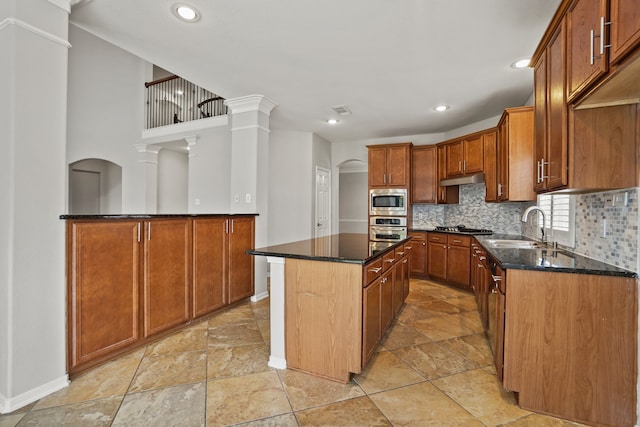 kitchen with dark stone counters, stainless steel appliances, decorative columns, a kitchen island, and sink