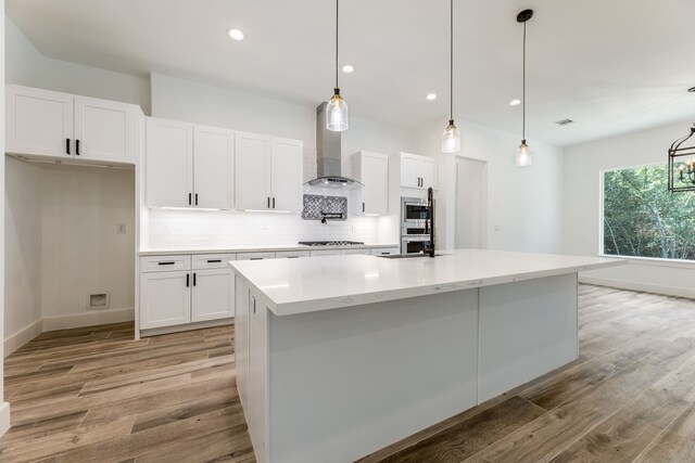 kitchen with white cabinetry, wall chimney exhaust hood, an island with sink, decorative light fixtures, and appliances with stainless steel finishes