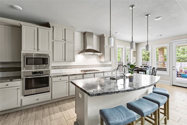 kitchen featuring decorative light fixtures, a kitchen island with sink, stainless steel appliances, wall chimney range hood, and light wood-type flooring