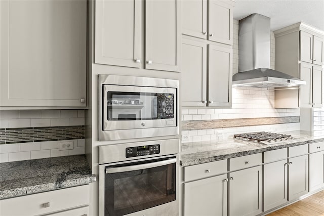 kitchen with wall chimney exhaust hood, backsplash, appliances with stainless steel finishes, white cabinetry, and light wood-type flooring