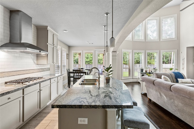 kitchen featuring a kitchen island with sink, stainless steel gas cooktop, decorative light fixtures, sink, and wall chimney exhaust hood