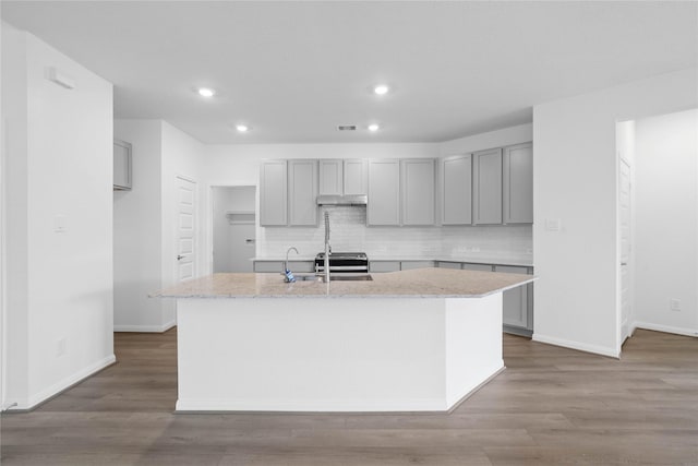 kitchen featuring light stone countertops, backsplash, a center island with sink, hardwood / wood-style flooring, and gray cabinets