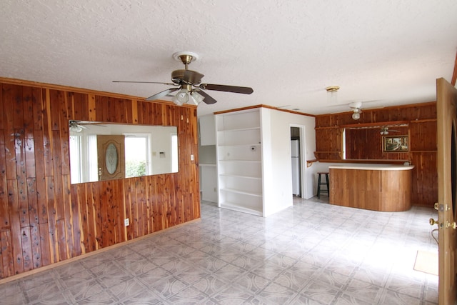 interior space with a textured ceiling, crown molding, kitchen peninsula, ceiling fan, and wooden walls