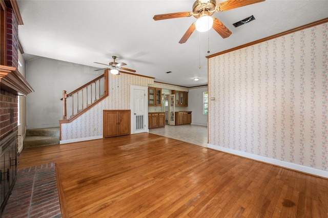 unfurnished living room featuring hardwood / wood-style floors, a ceiling fan, wallpapered walls, ornamental molding, and stairs