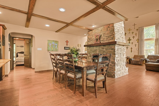 dining room featuring light wood-type flooring, coffered ceiling, beam ceiling, and a stone fireplace