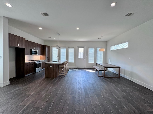 kitchen featuring stainless steel appliances, dark hardwood / wood-style flooring, an island with sink, hanging light fixtures, and dark brown cabinetry