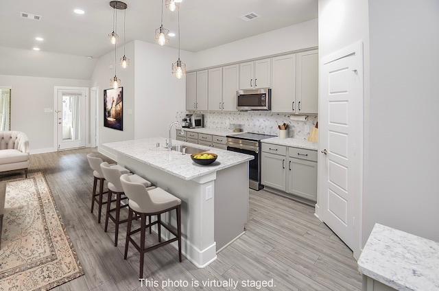 kitchen featuring stainless steel appliances, lofted ceiling, decorative light fixtures, a center island with sink, and light wood-type flooring