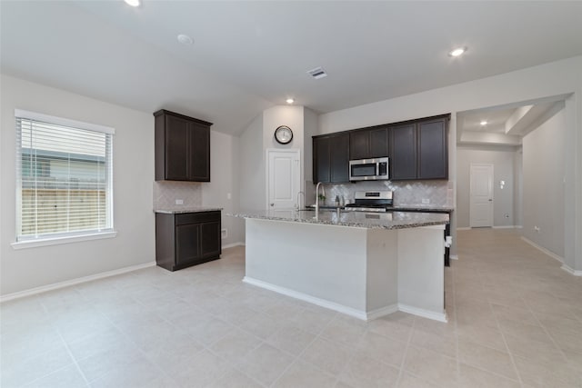 kitchen with backsplash, appliances with stainless steel finishes, a kitchen island with sink, light stone counters, and dark brown cabinetry