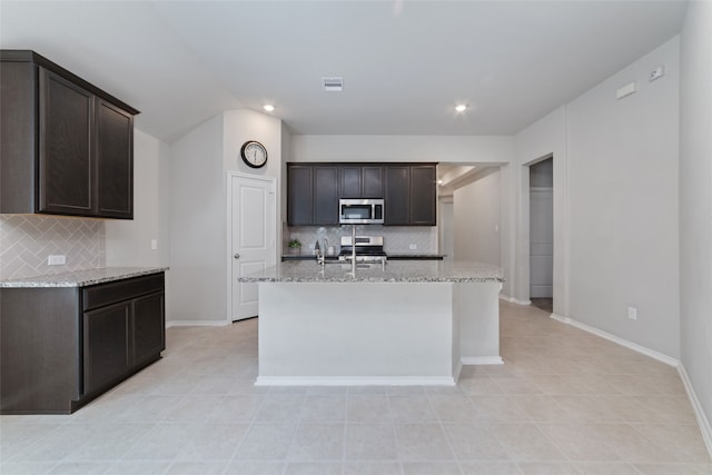 kitchen featuring light stone countertops, dark brown cabinets, appliances with stainless steel finishes, tasteful backsplash, and an island with sink