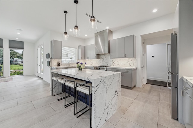 kitchen featuring a kitchen island, gray cabinetry, a kitchen breakfast bar, wall chimney exhaust hood, and light stone counters