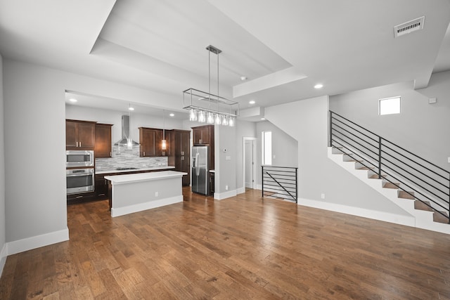 kitchen with appliances with stainless steel finishes, hanging light fixtures, dark wood-type flooring, wall chimney exhaust hood, and a center island