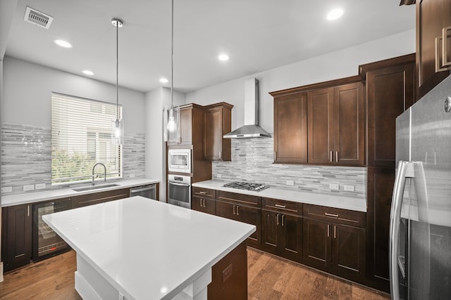 kitchen with wood-type flooring, beverage cooler, a kitchen island, wall chimney range hood, and appliances with stainless steel finishes