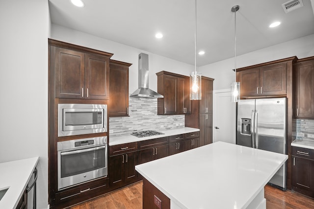 kitchen featuring pendant lighting, hardwood / wood-style flooring, a kitchen island, wall chimney range hood, and appliances with stainless steel finishes