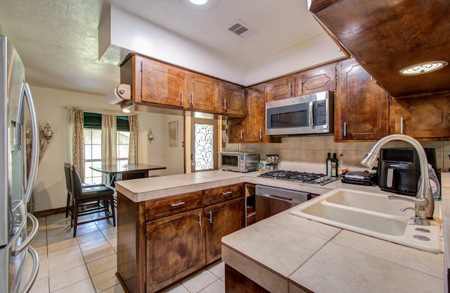 kitchen with appliances with stainless steel finishes, light tile patterned floors, sink, tile countertops, and kitchen peninsula