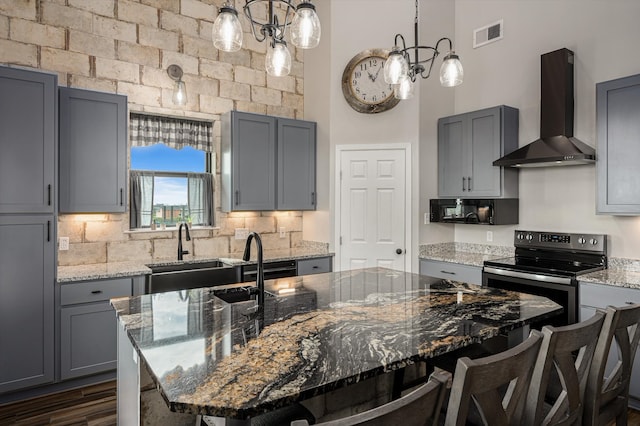 kitchen with wall chimney exhaust hood, stainless steel range with electric stovetop, light stone counters, and a kitchen island with sink