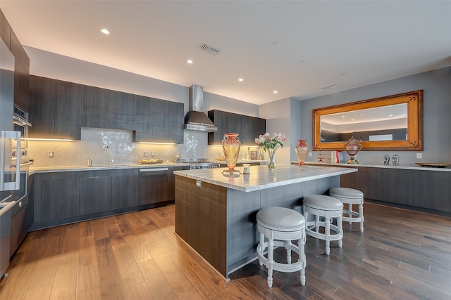 kitchen featuring backsplash, wood-type flooring, a center island, a breakfast bar area, and wall chimney range hood