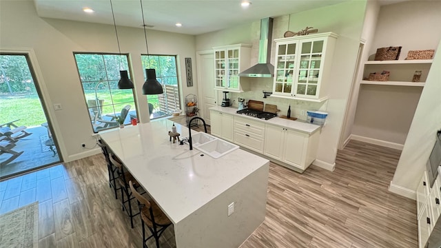 kitchen featuring light stone countertops, wall chimney exhaust hood, a center island with sink, pendant lighting, and white cabinetry