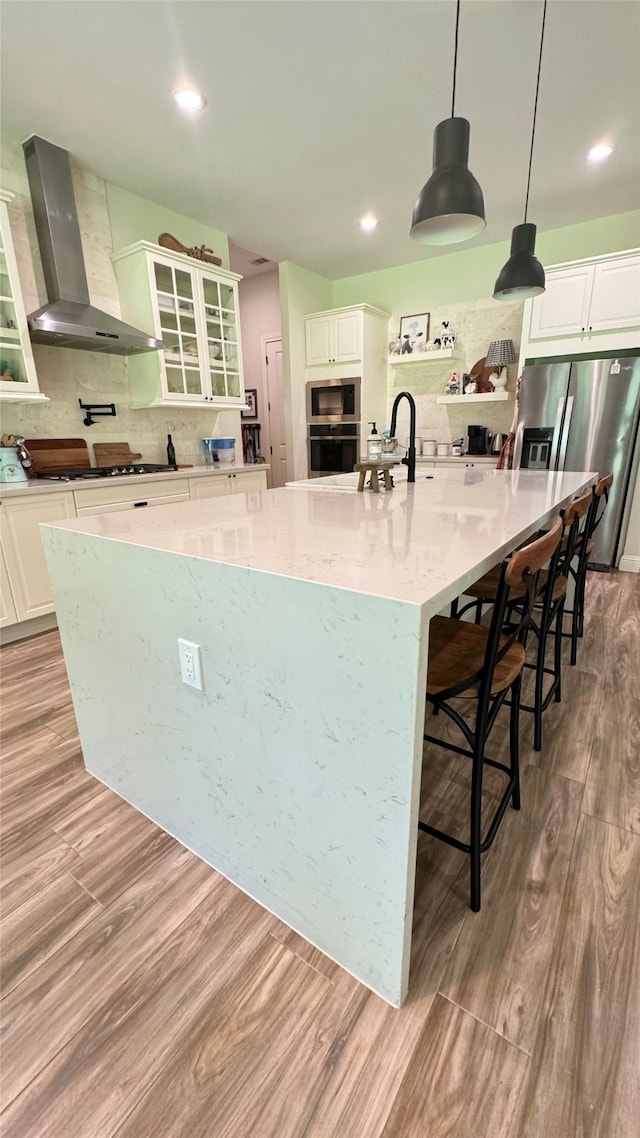 kitchen with appliances with stainless steel finishes, light hardwood / wood-style floors, hanging light fixtures, a large island, and wall chimney range hood