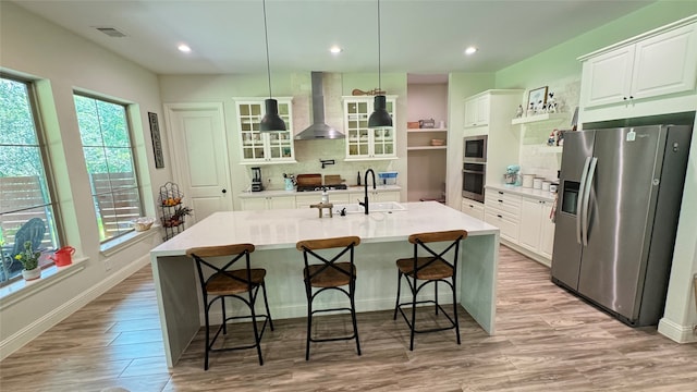 kitchen with white cabinetry, an island with sink, hanging light fixtures, appliances with stainless steel finishes, and wall chimney range hood