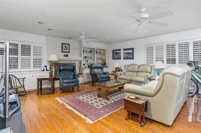 living room featuring light wood-type flooring, built in shelves, ceiling fan, and a brick fireplace