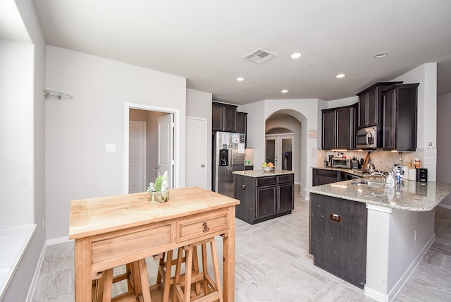 kitchen featuring dark brown cabinetry, appliances with stainless steel finishes, light stone counters, a sink, and arched walkways