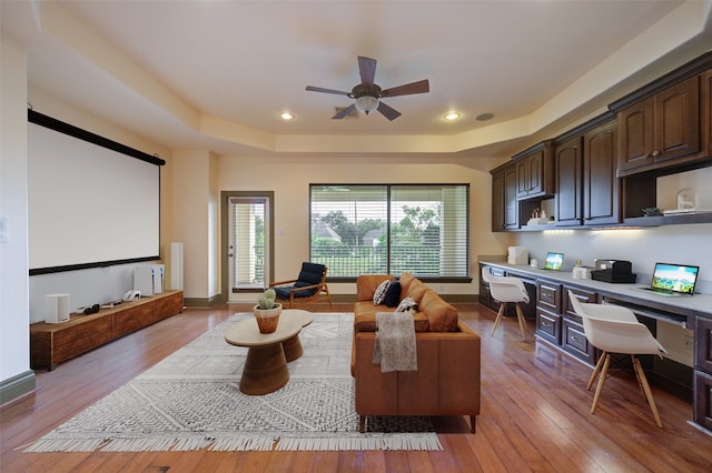 cinema featuring built in desk, ceiling fan, a tray ceiling, and light hardwood / wood-style flooring