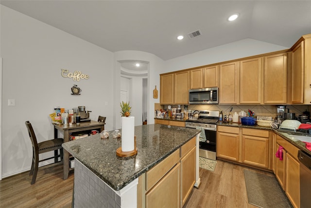 kitchen featuring light wood-type flooring, tasteful backsplash, stainless steel appliances, a center island, and dark stone counters