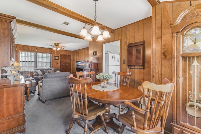 dining room with carpet floors, beamed ceiling, ceiling fan with notable chandelier, and wood walls