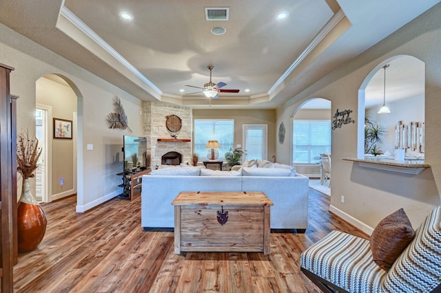 living room with a tray ceiling, hardwood / wood-style flooring, a stone fireplace, ornamental molding, and ceiling fan