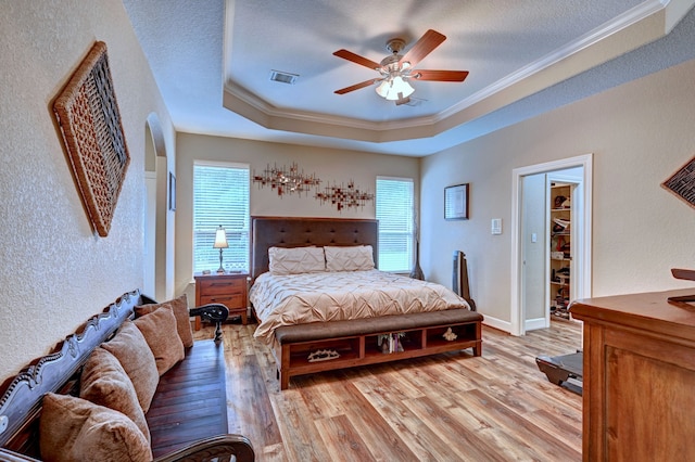 bedroom with light wood-type flooring, a tray ceiling, ceiling fan, a walk in closet, and ornamental molding