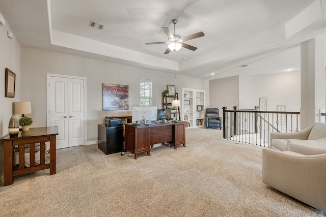 carpeted home office with ceiling fan and a raised ceiling