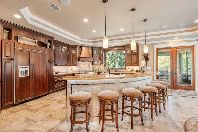 kitchen featuring premium range hood, light stone countertops, a kitchen island with sink, and a raised ceiling