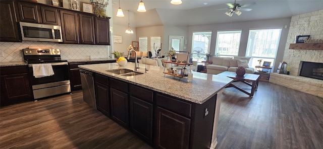 kitchen with appliances with stainless steel finishes, an island with sink, a stone fireplace, dark hardwood / wood-style flooring, and ceiling fan