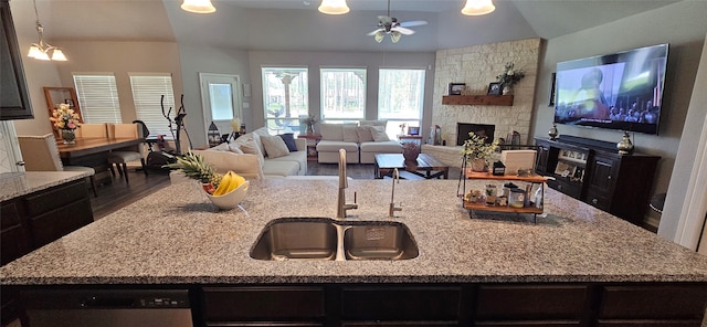 kitchen with dark hardwood / wood-style floors, a kitchen island with sink, ceiling fan with notable chandelier, a stone fireplace, and stainless steel dishwasher